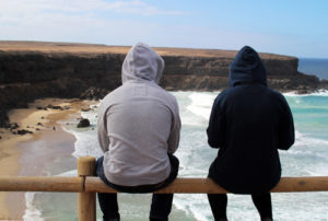 Surfer couple sitting on a fence wearing hoodies overlooking the waves hitting the beach